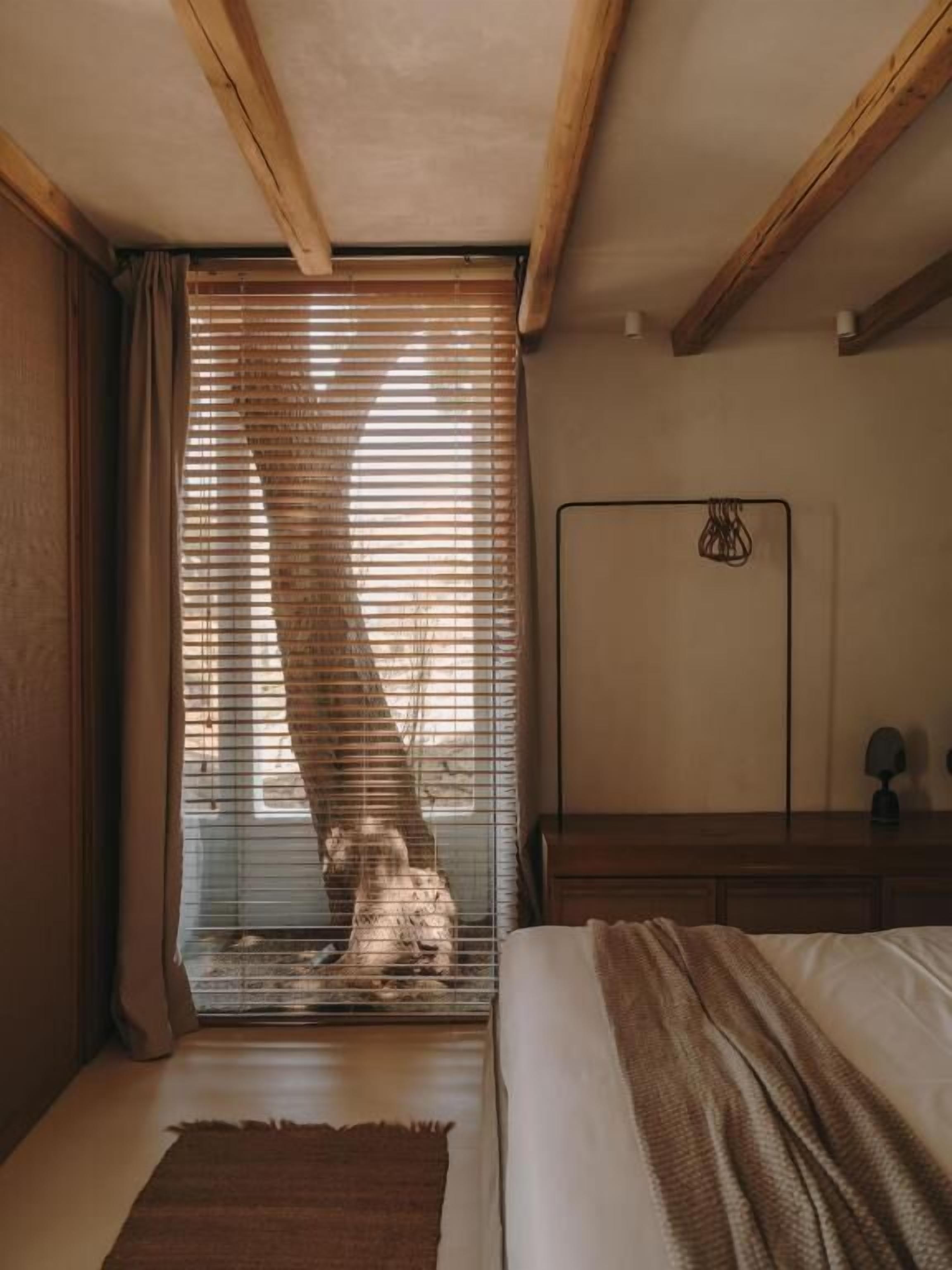A bedroom with a bed, a wooden headboard, a bedside table, a floor lamp, and a window with blinds.