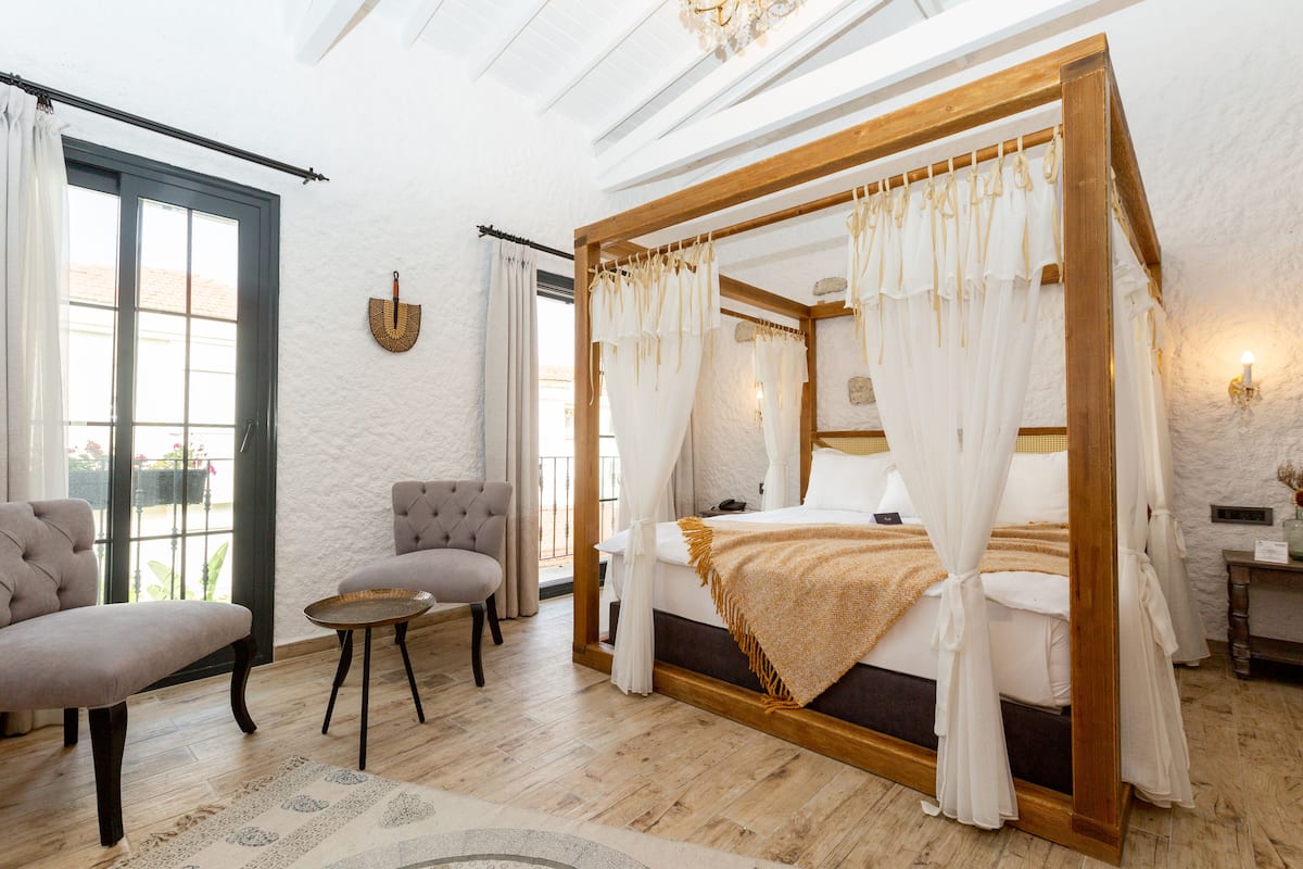 A bedroom with a four-poster bed, a sitting area with a chair and table, and a view through a window.