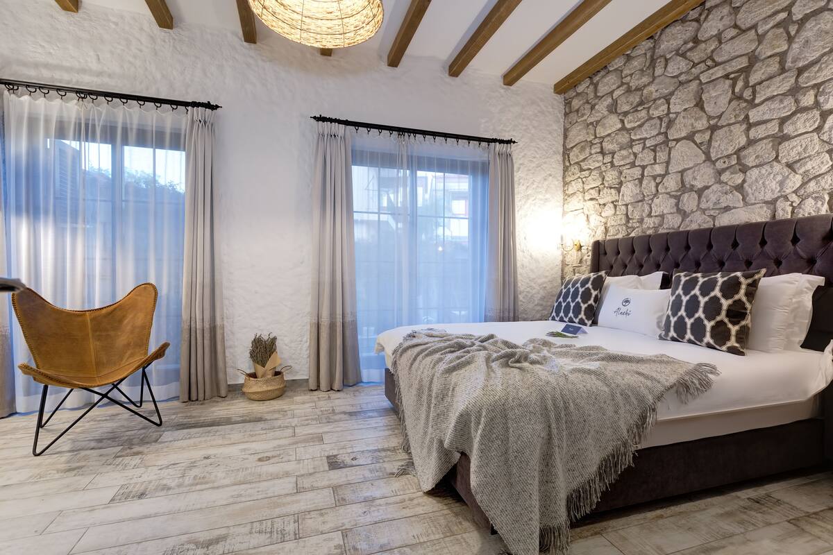 A bedroom with a stone wall, a bed with patterned pillows, a wooden chair, and a large window with curtains.