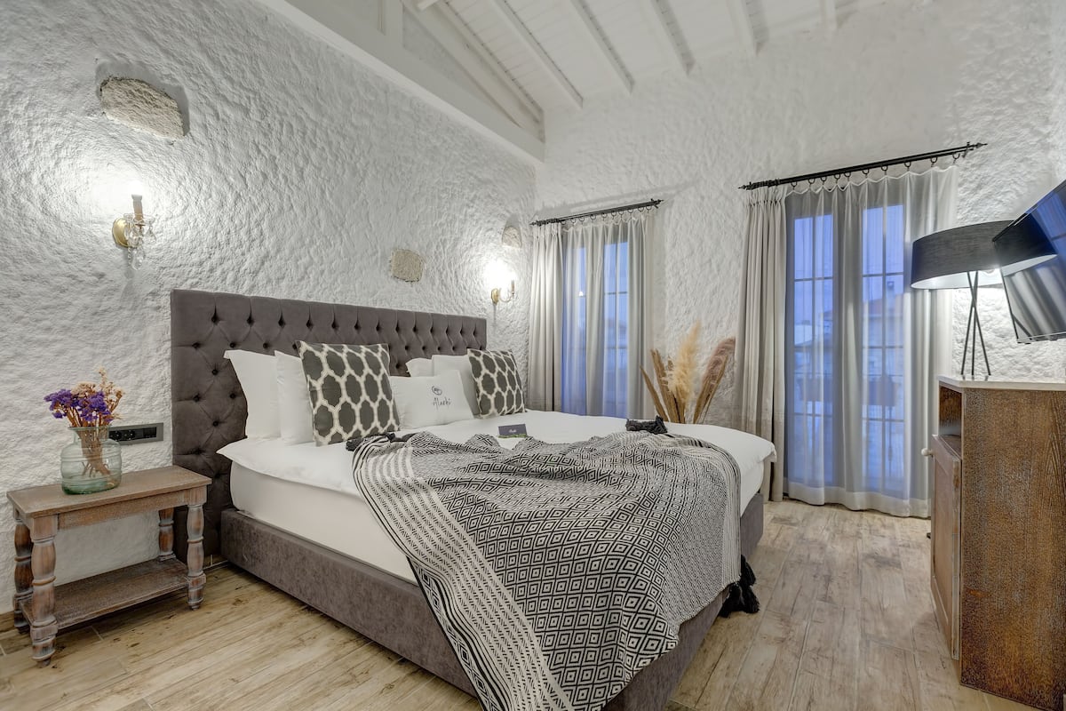 A bedroom with a tufted headboard, a bedside table with a vase of flowers, a patterned throw blanket, and a lamp on a wooden cabinet.