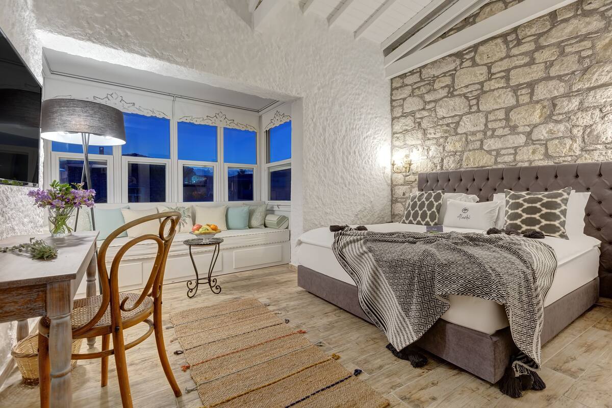 A bedroom with a stone wall, a large bed with patterned pillows, a window with a view, and a dining area with a table and chair.