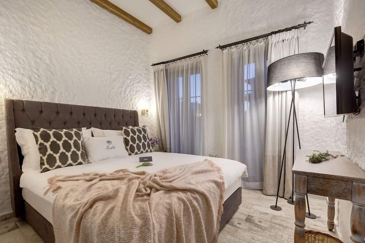 A bedroom with a tufted headboard, a bedside table, a lamp, and a window with curtains.