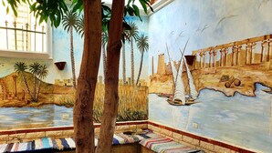 A room with a mural depicting a coastal scene, a bench with colorful rugs, and a tree in the foreground. - Spring Hotel Luxor (Luxor)