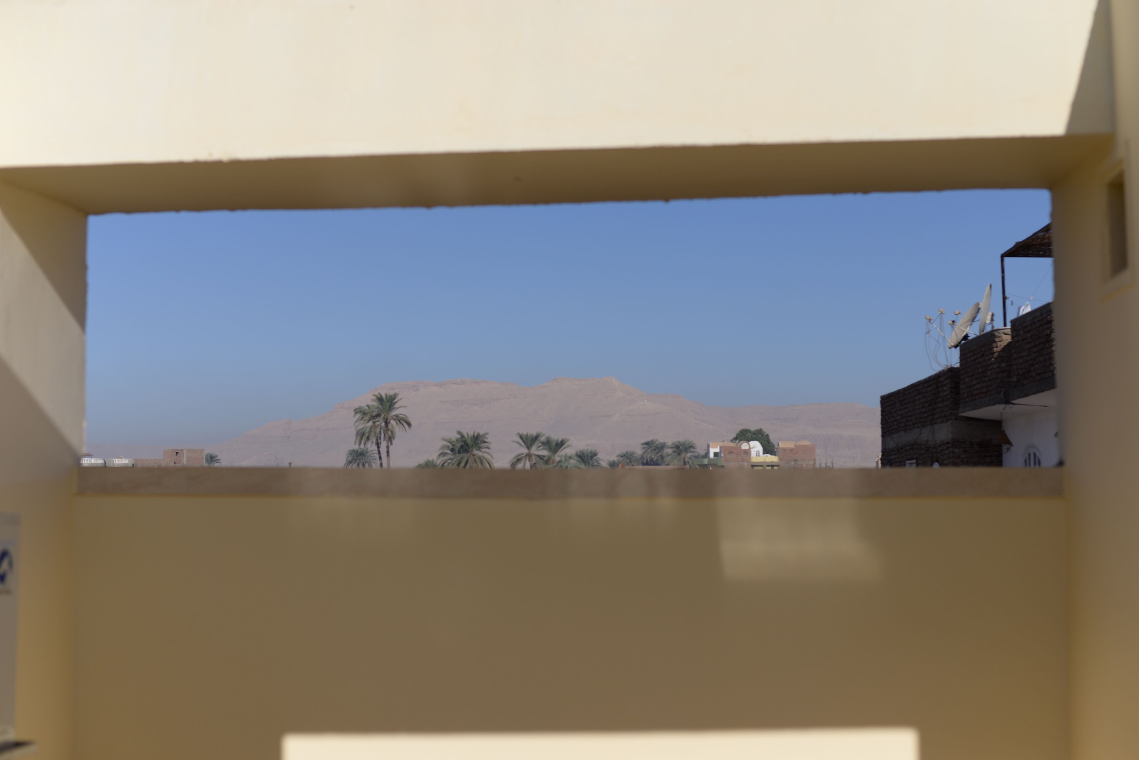 Blick durch ein Fenster mit einem Berg im Hintergrund, Palmen und einem Gebäude mit Satellitenschüsseln.