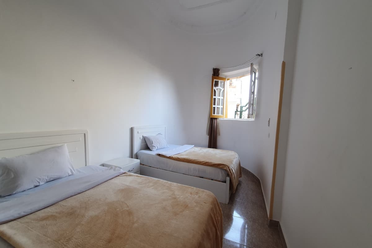 A small, bright room with two single beds, a window with a view of the outside, and a white ceiling.