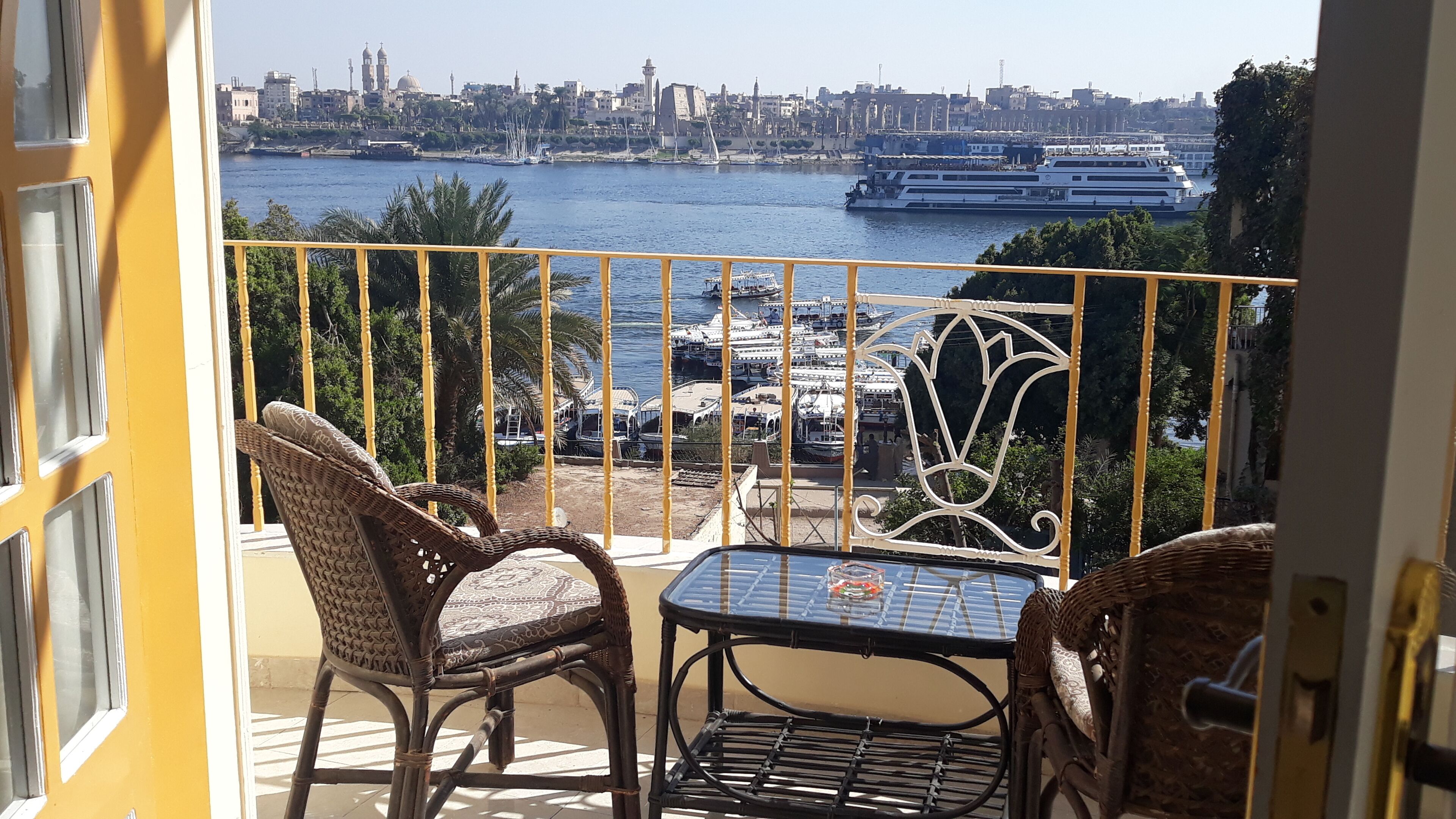 Un balcón con sillas de mimbre y una mesa, con vista a un río con barcos y un paisaje urbano de fondo.