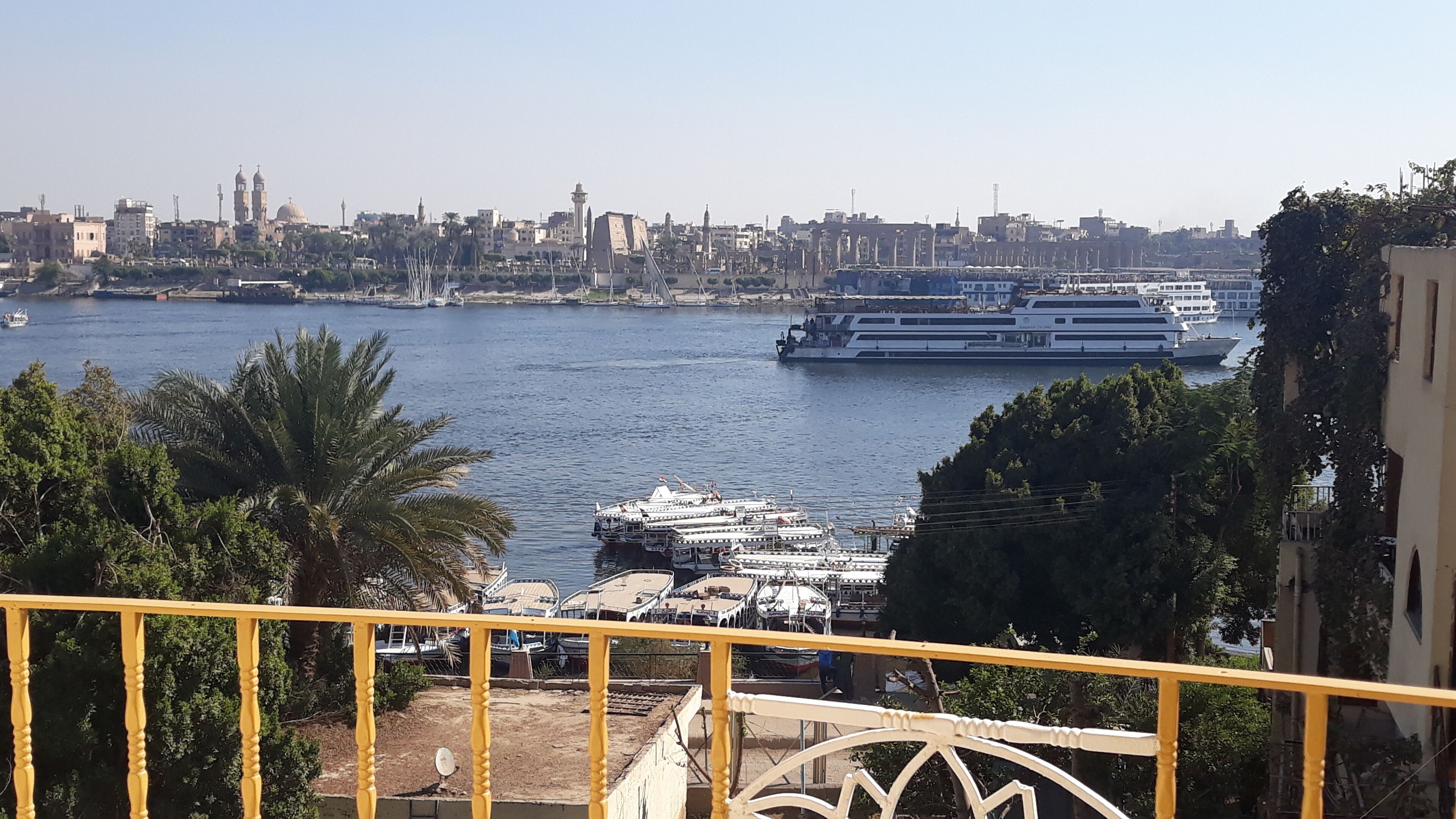 Un río con barcos, el perfil urbano de una ciudad y una baranda amarilla al frente.