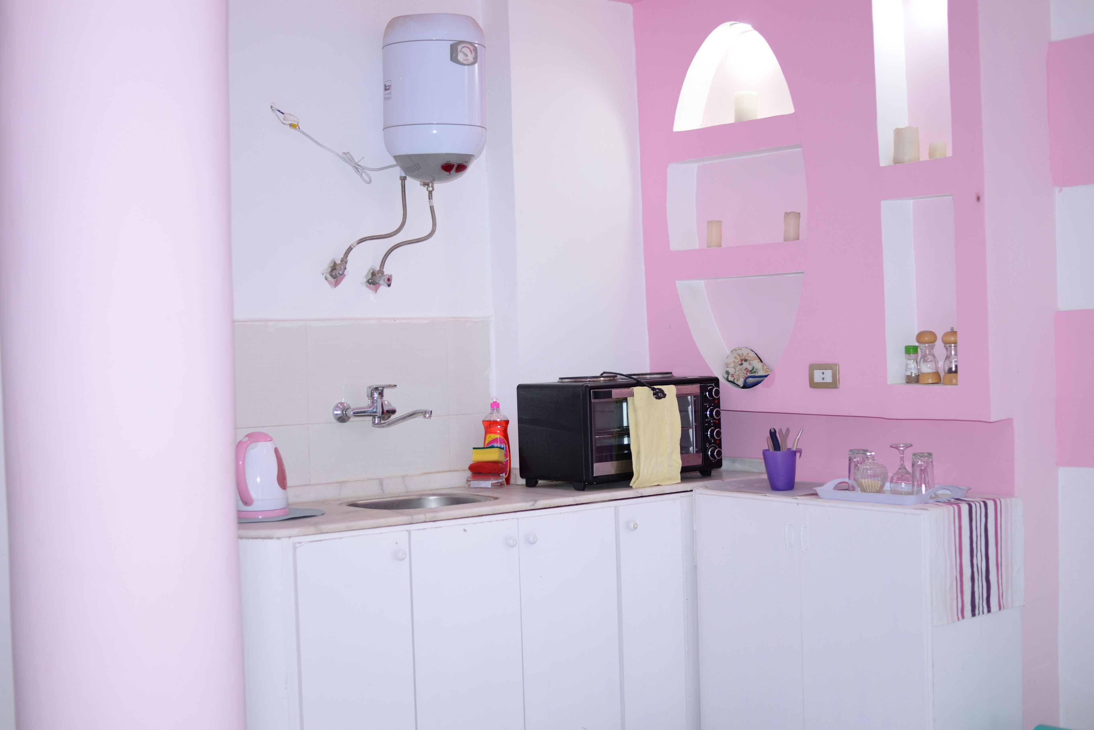 A kitchen with white cabinets, a pink wall, a sink, a kettle, a toaster, and a water heater.