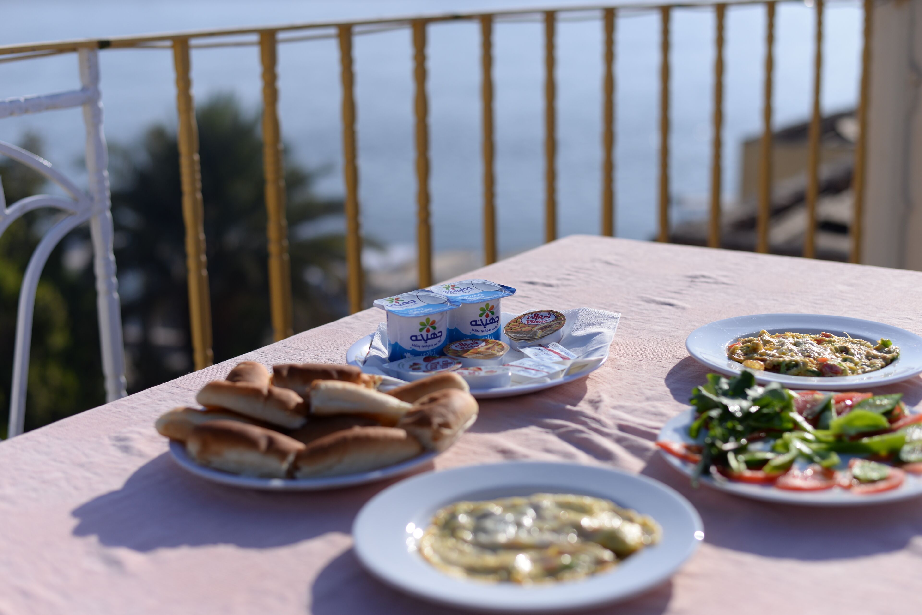 A table with food items including bread, yogurt, and a salad, set on a balcony with a railing and a view of the sea.