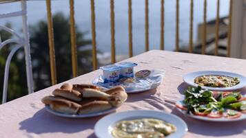 Una mesa con alimentos como pan, yogur y ensalada, ubicada en un balcón con barandilla y vistas al mar.