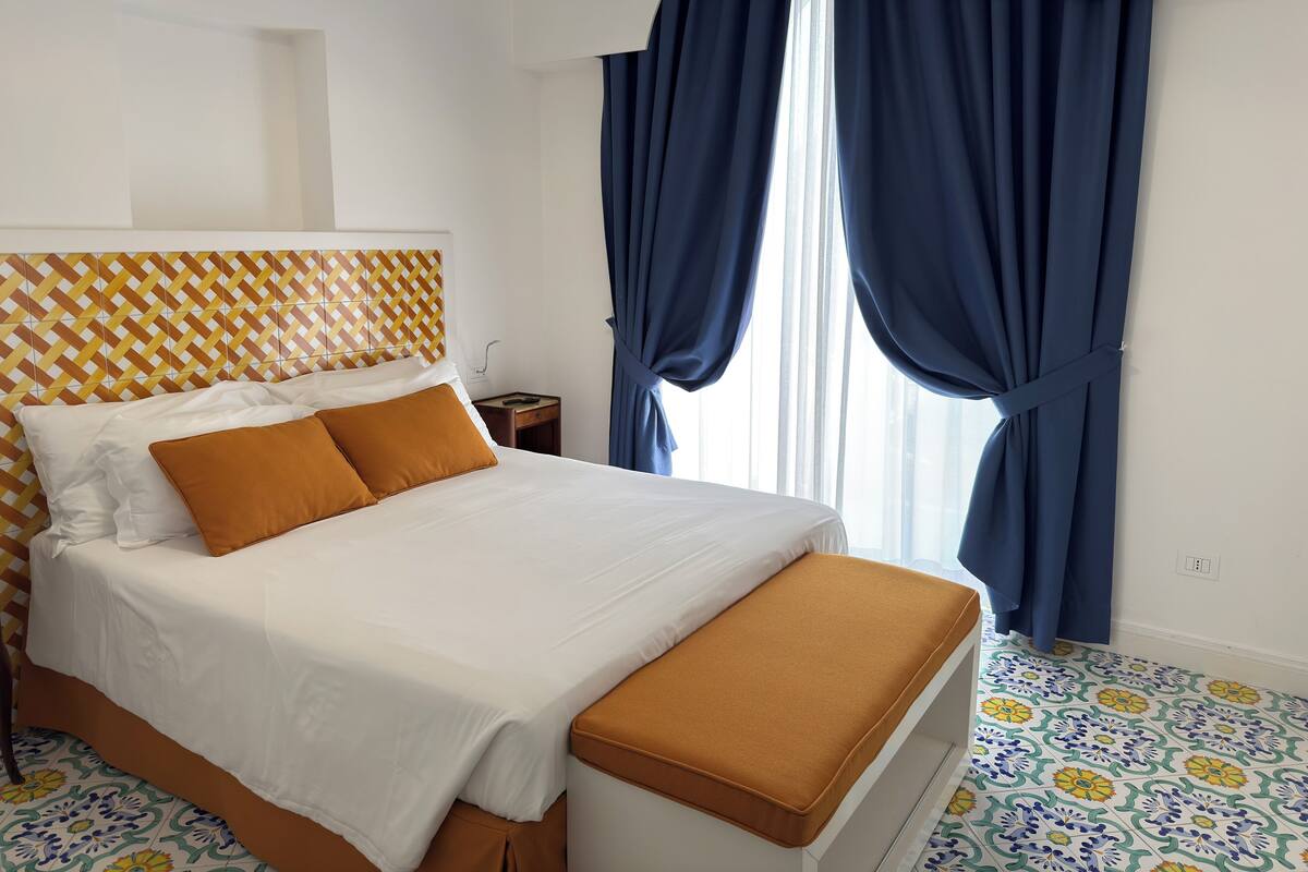 A neatly made bed with white and mustard bedding, a patterned headboard, and a wooden nightstand with a remote control.