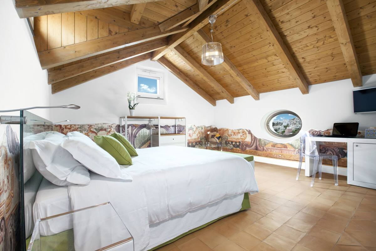 A bedroom with a wooden ceiling, a bed with white bedding, a desk with a chair, and a view through a window.