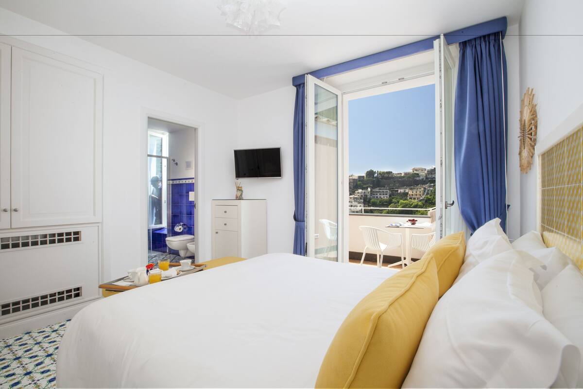 A hotel room with a large bed, a television, a balcony with a view of the city, and a bathroom with blue tiles.