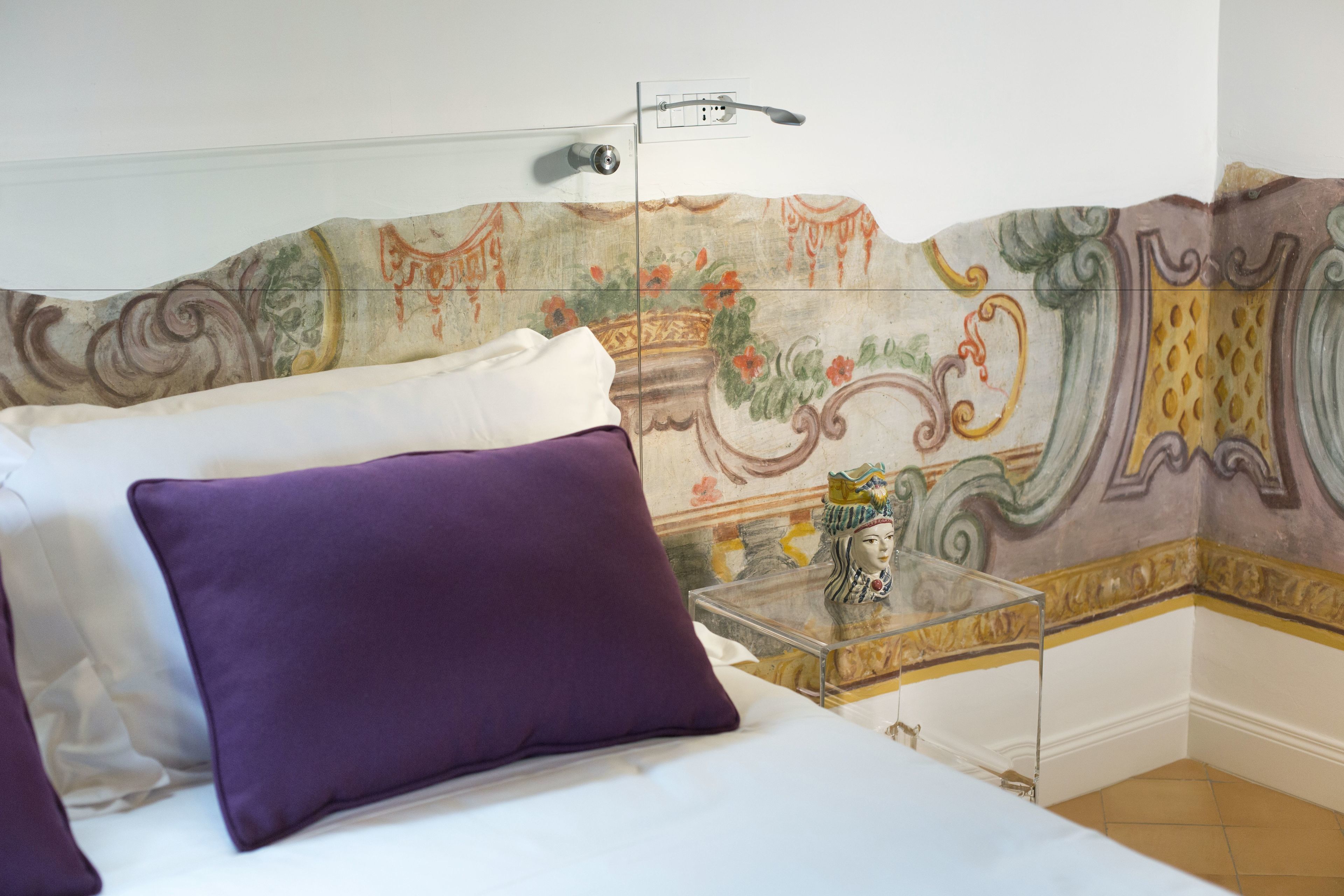 A bedroom with a bed, purple pillows, a clear glass side table, and a decorative wall with a floral pattern.