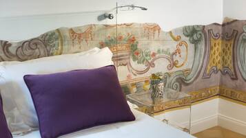 Un dormitorio con una cama, almohadas moradas, una mesita de noche de vidrio transparente y una pared decorativa con estampado floral.