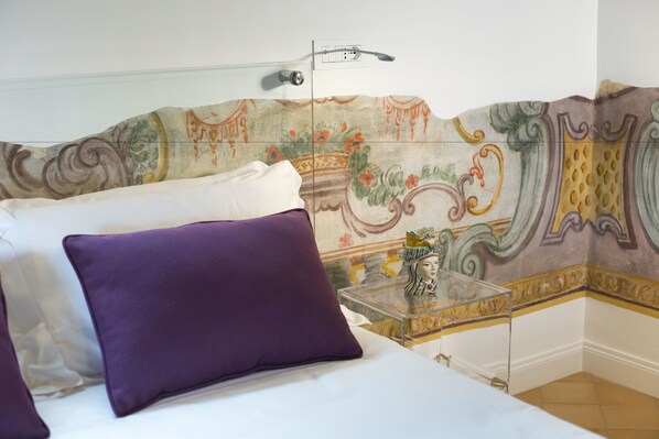 Un dormitorio con una cama, almohadas moradas, una mesita de noche de vidrio transparente y una pared decorativa con estampado floral.