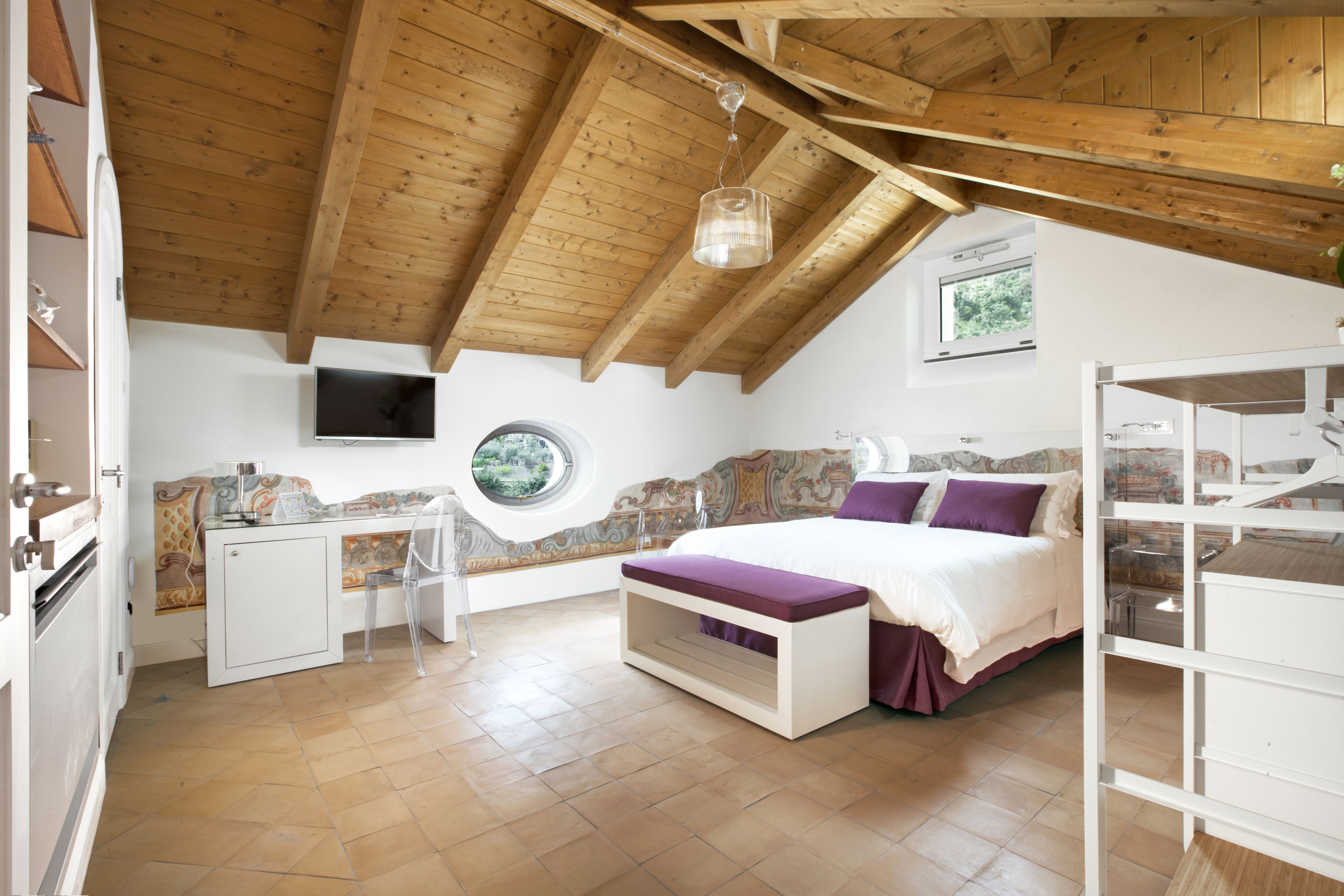 Een slaapkamer met een houten plafond, een bed met een paars dekbed, een bureau met een stoel en een raam met uitzicht op groen.