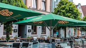 Terrasse mit grünen Sonnenschirmen, Holztischen und -stühlen, vor einem Gebäude mit großen Fenstern und einem Baum.