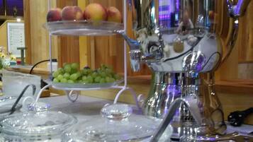 A buffet setup with a fruit display, a beverage dispenser, and glass containers with utensils.