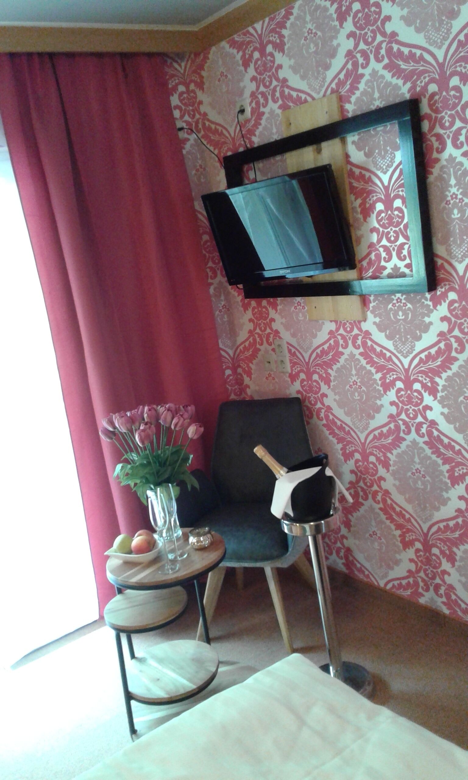 A room with a TV mounted on the wall, a chair, a small table with a vase of flowers, and a window with red curtains.