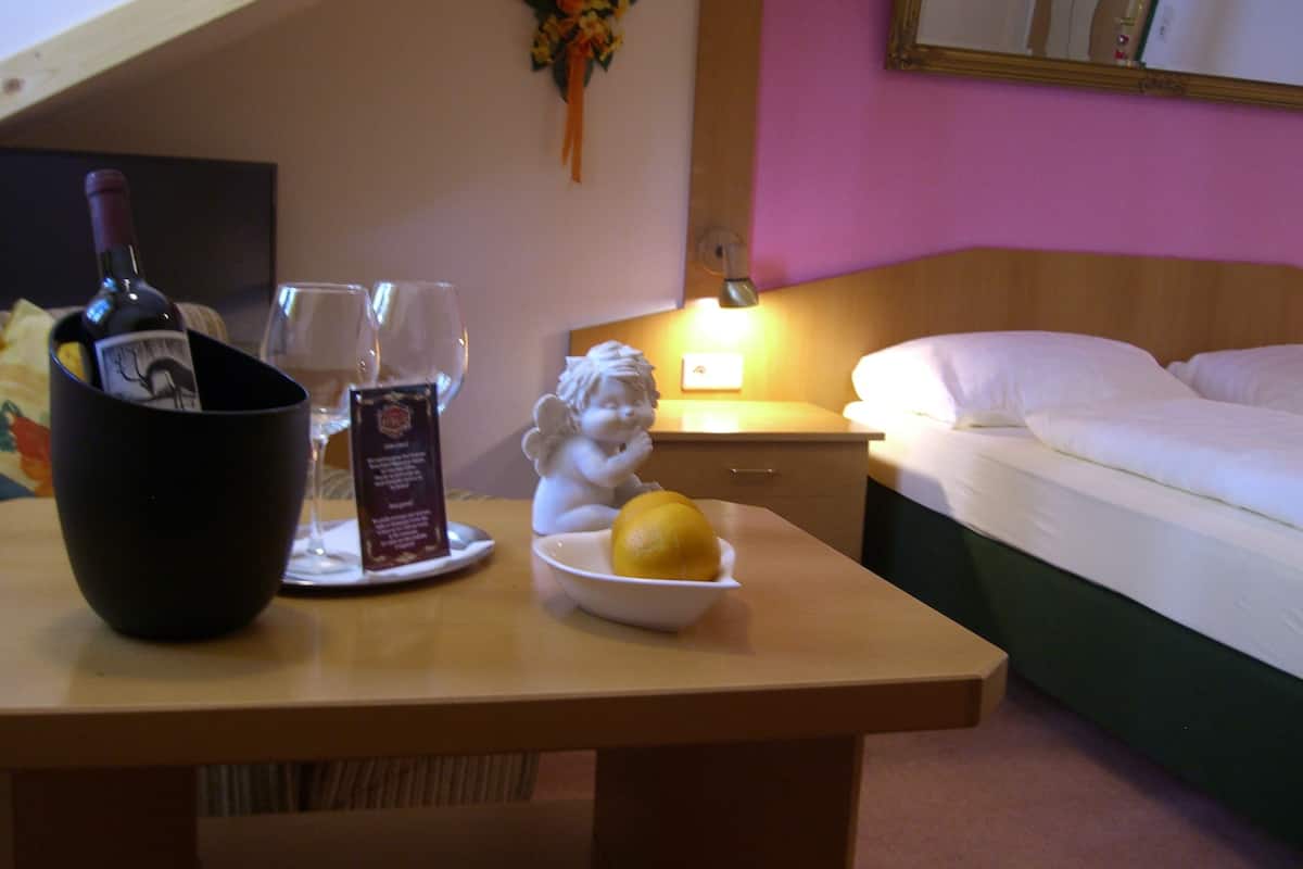 A hotel room with a bed, a nightstand, a table with a wine bottle, glasses, a menu, and a fruit bowl.