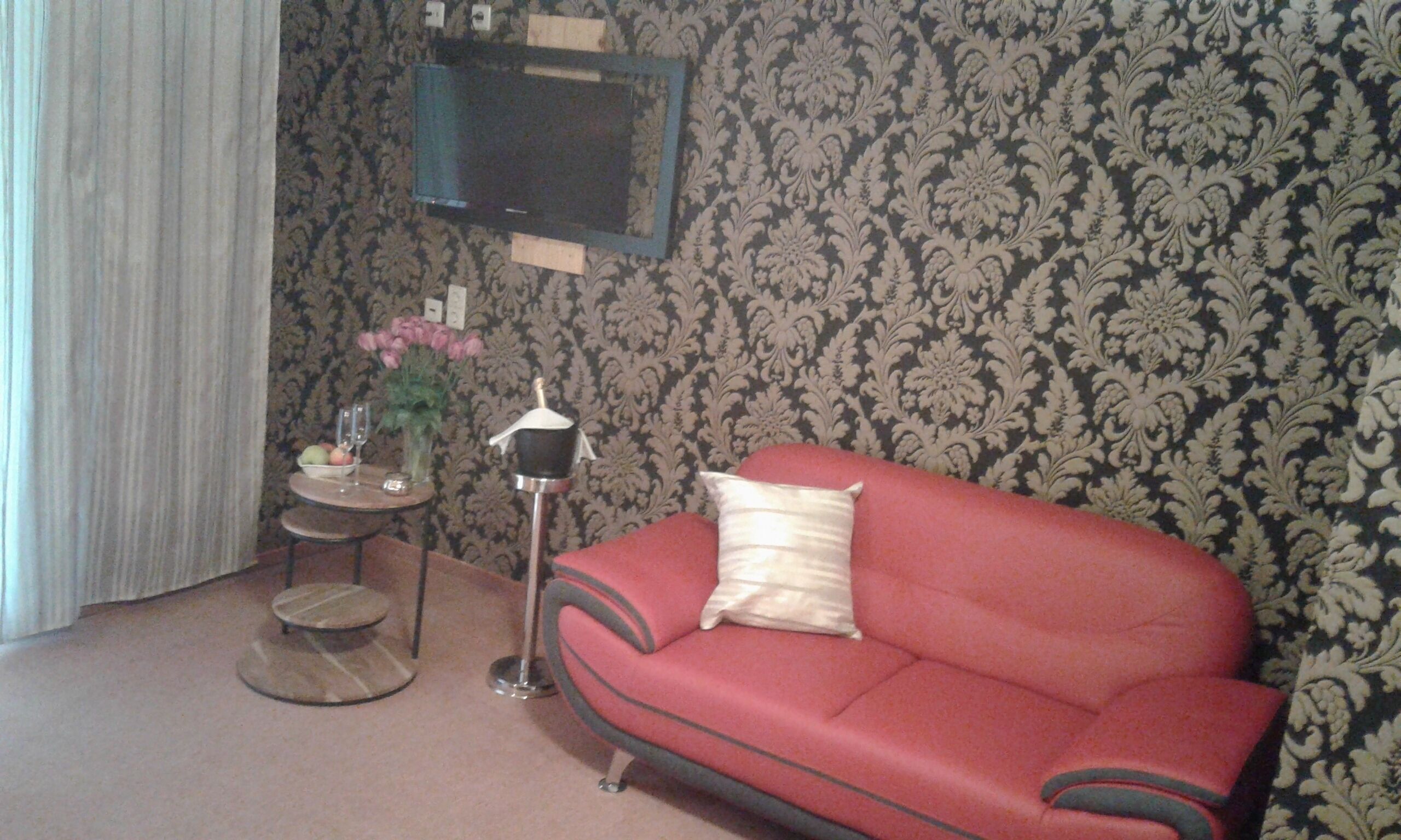 A living room with a red sofa, a small round table with a vase of flowers, and a television mounted on the wall.