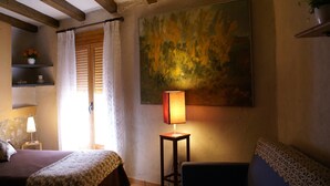 A room with a bed, a chair, a table with a lamp, a painting on the wall, and a window with curtains.