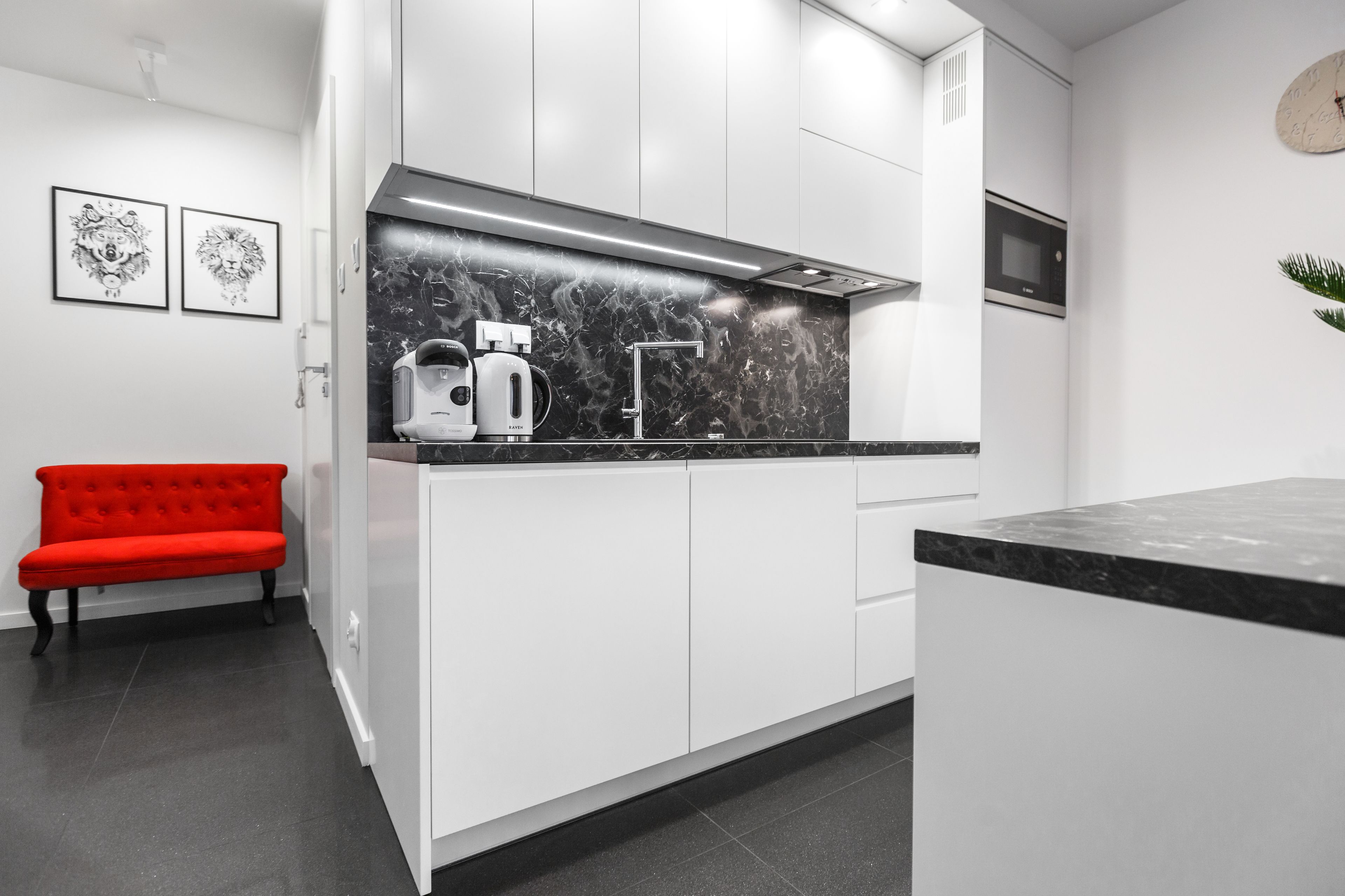 Une cuisine moderne avec des meubles blancs, un plan de travail en marbre noir, un banc rouge et un four à micro-ondes.