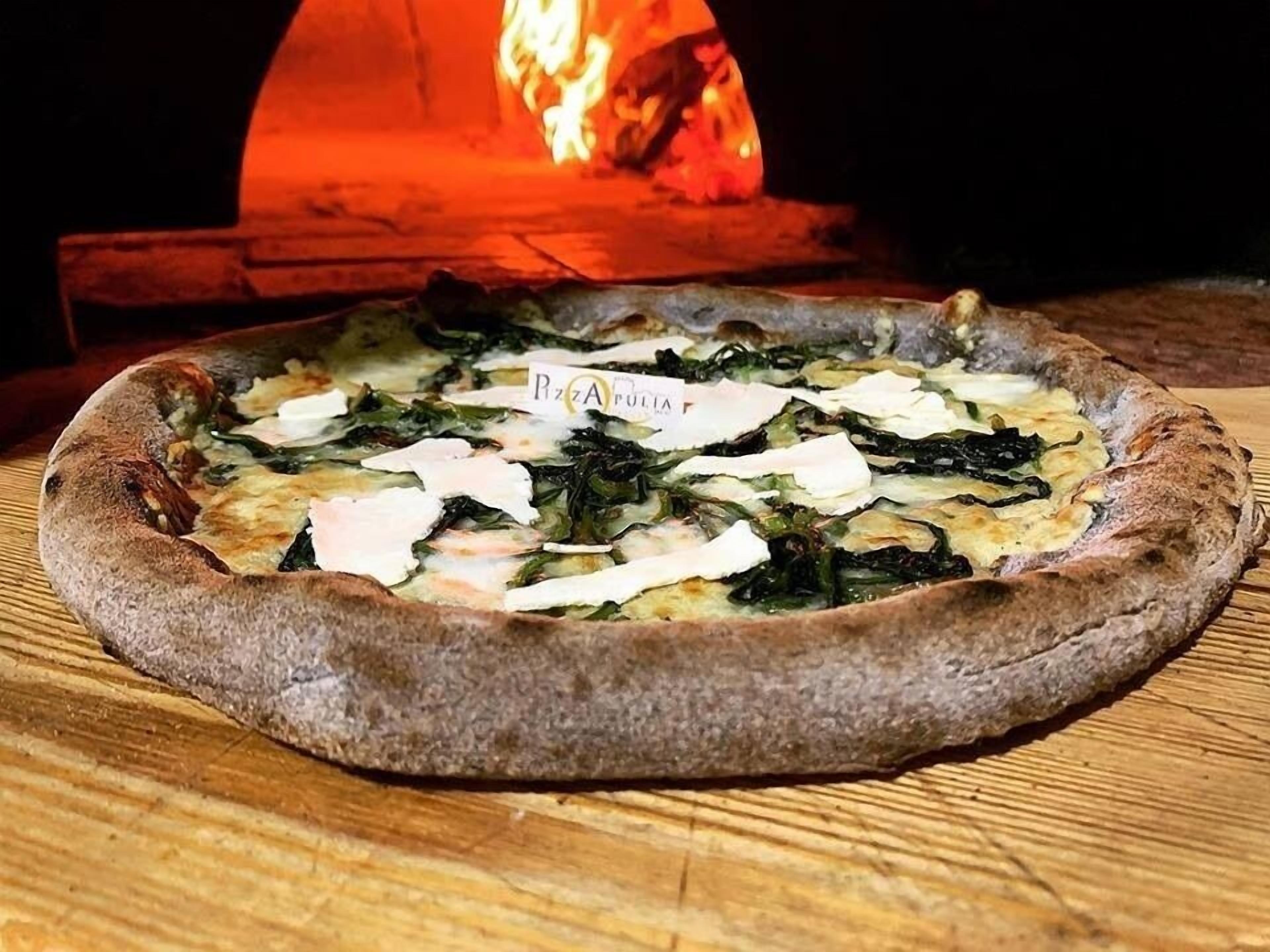 Pizza con formaggio e verdure su un tavolo di legno, con un forno a legna sullo sfondo.