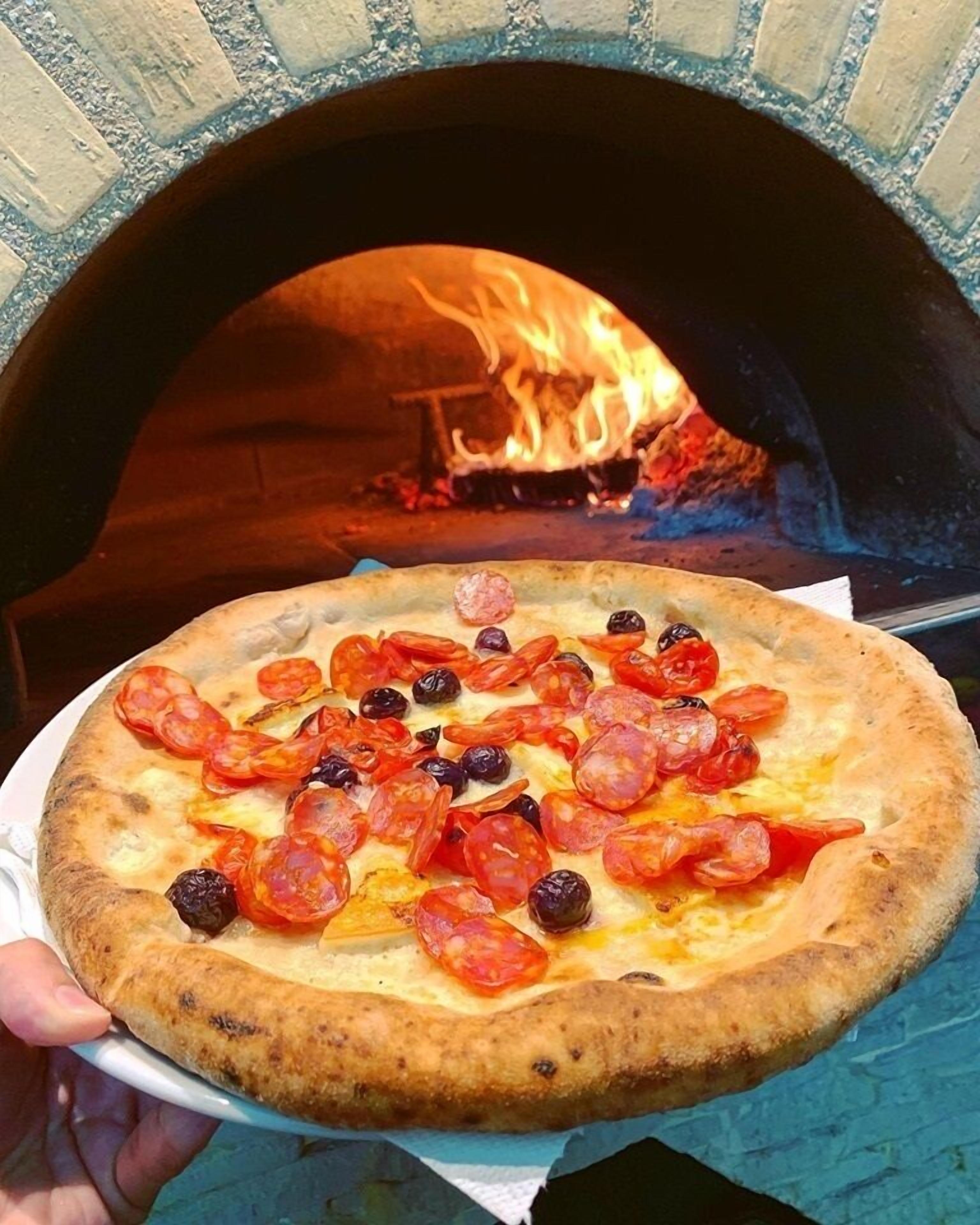 A pizza with pepperoni and olives being held in front of a brick oven with a fire.
