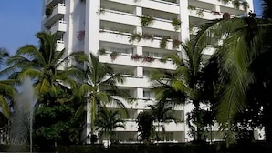 A tall, white hotel with balconies adorned with plants, surrounded by palm trees and a body of water. - Condo M Principe (Ixtapa)