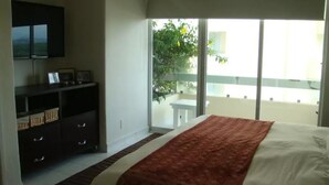 A hotel room with a bed, a TV mounted on the wall, a small table, and a balcony with a view of greenery. - Condo M Principe (Ixtapa)