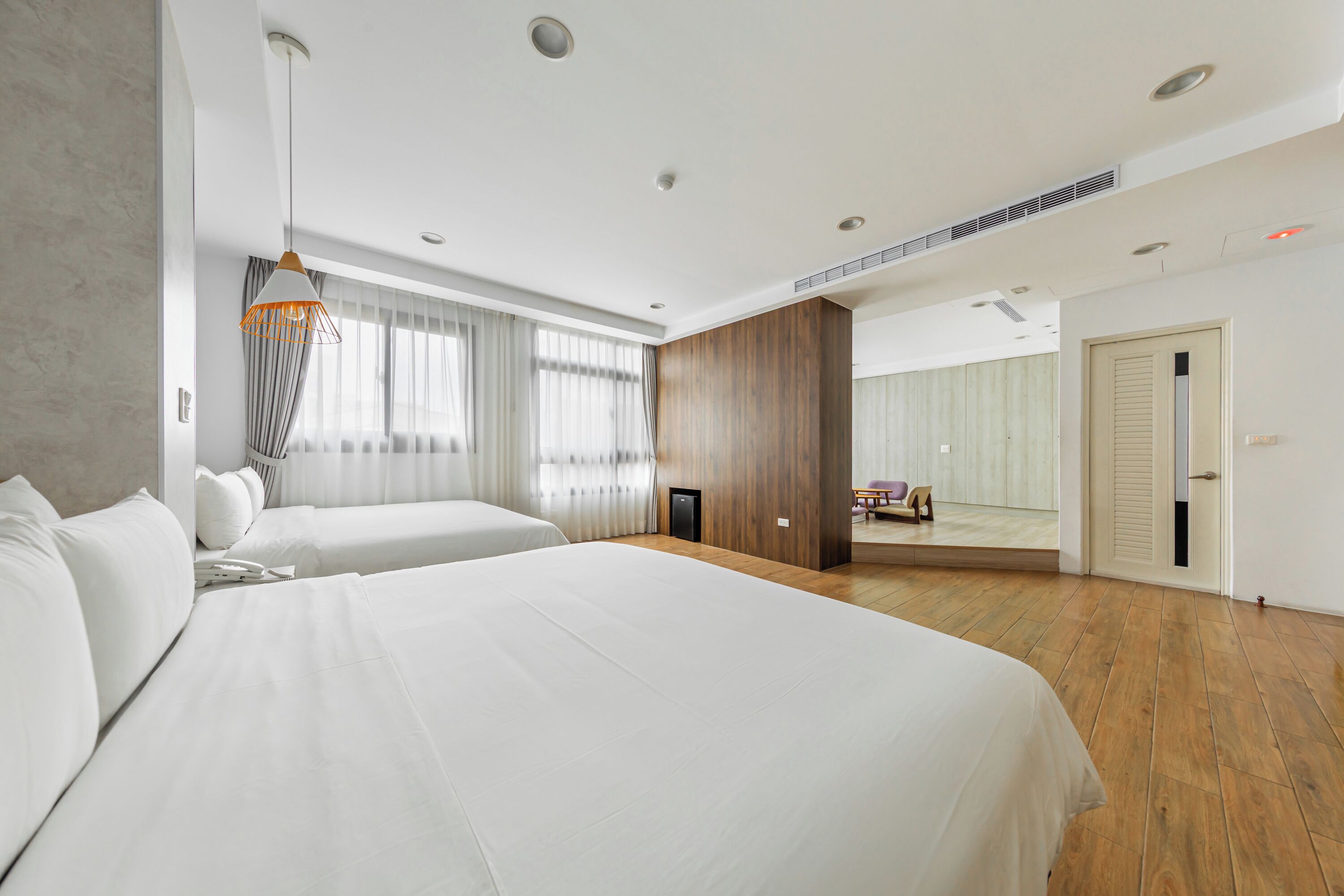 Quarto de hotel moderno com duas camas grandes, piso de madeira, uma janela ampla com cortinas e vista para uma varanda.