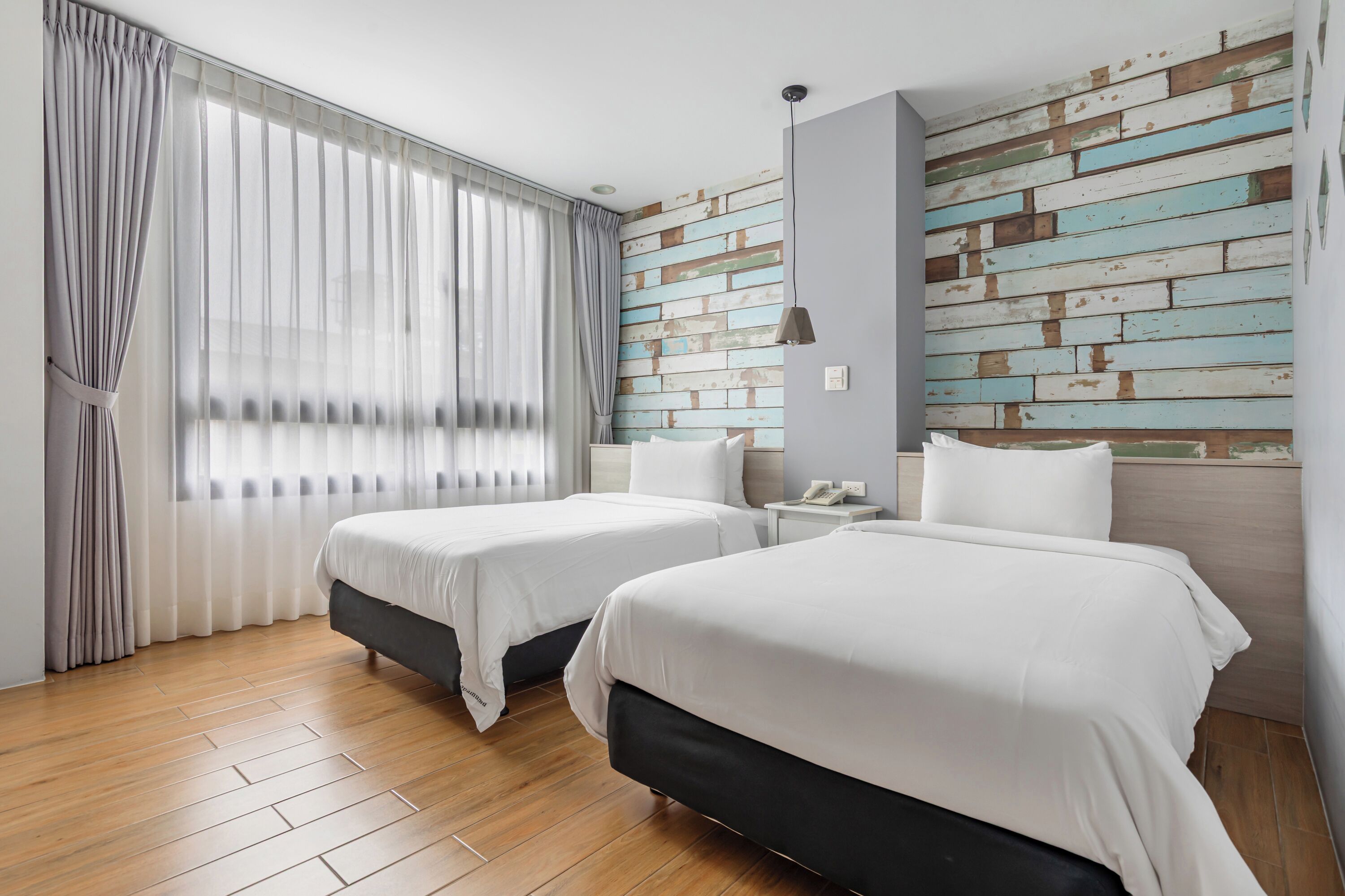 A hotel room with two beds, wooden flooring, a brick-patterned wall, and a window with sheer curtains.