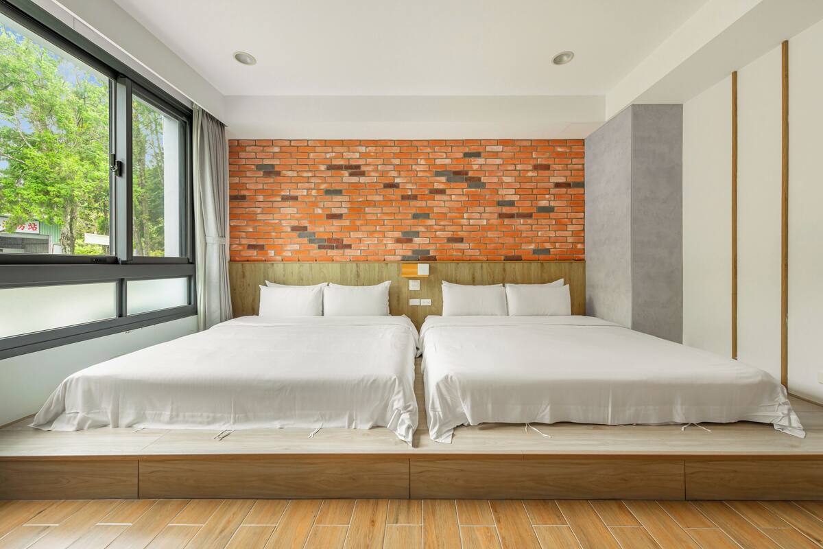 A modern hotel room with a brick-patterned headboard, two beds with white linens, a large window with curtains, and a wooden floor.