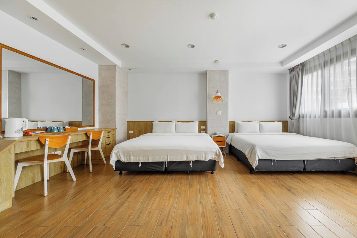 A hotel room with a wooden floor, two beds, a desk with chairs, a large mirror, and a window with curtains.