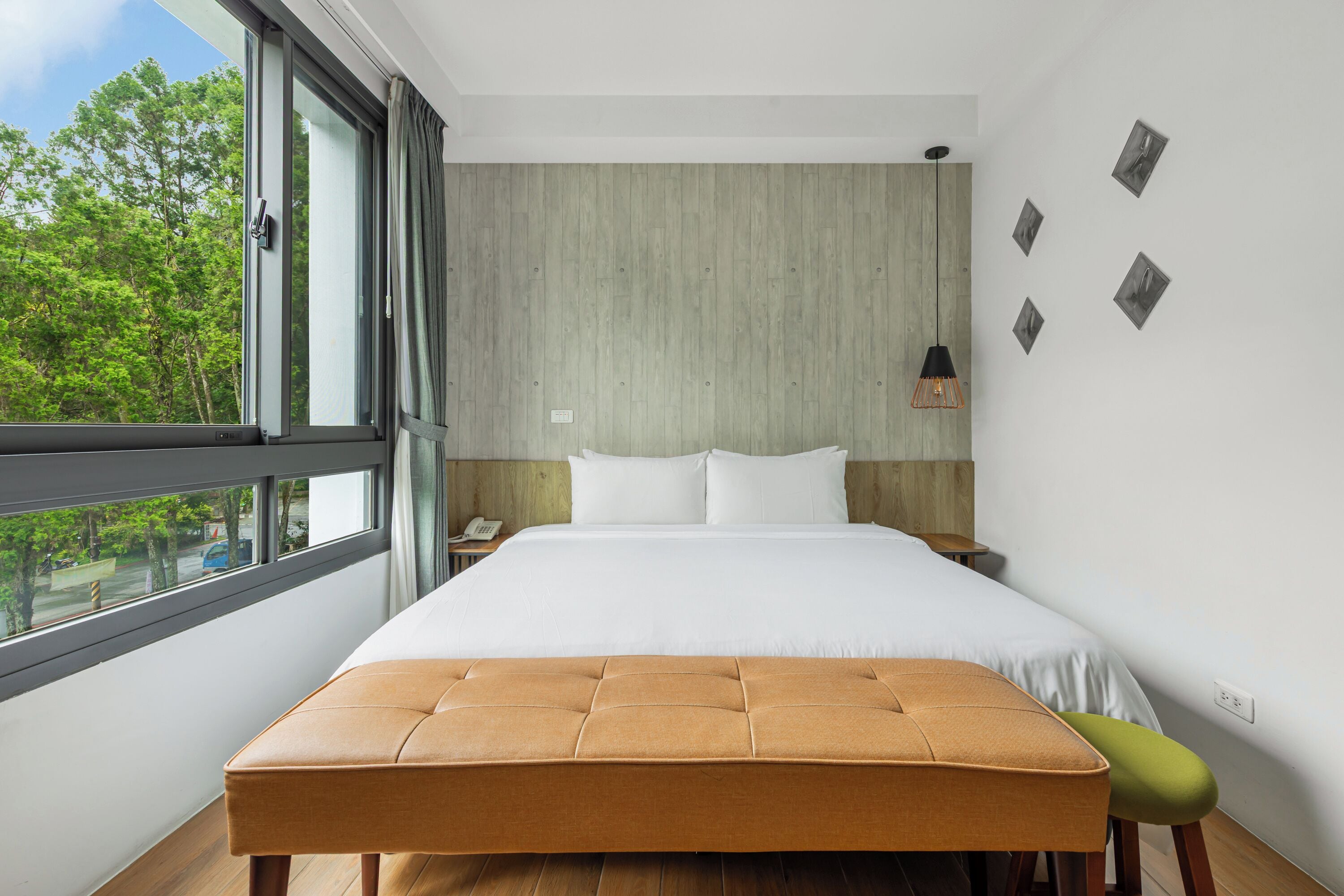 A modern bedroom with a large bed, a wooden bench, a nightstand, and a view of trees through a large window.