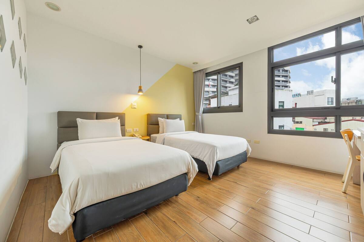 A hotel room with two beds, a large window, and a view of buildings.