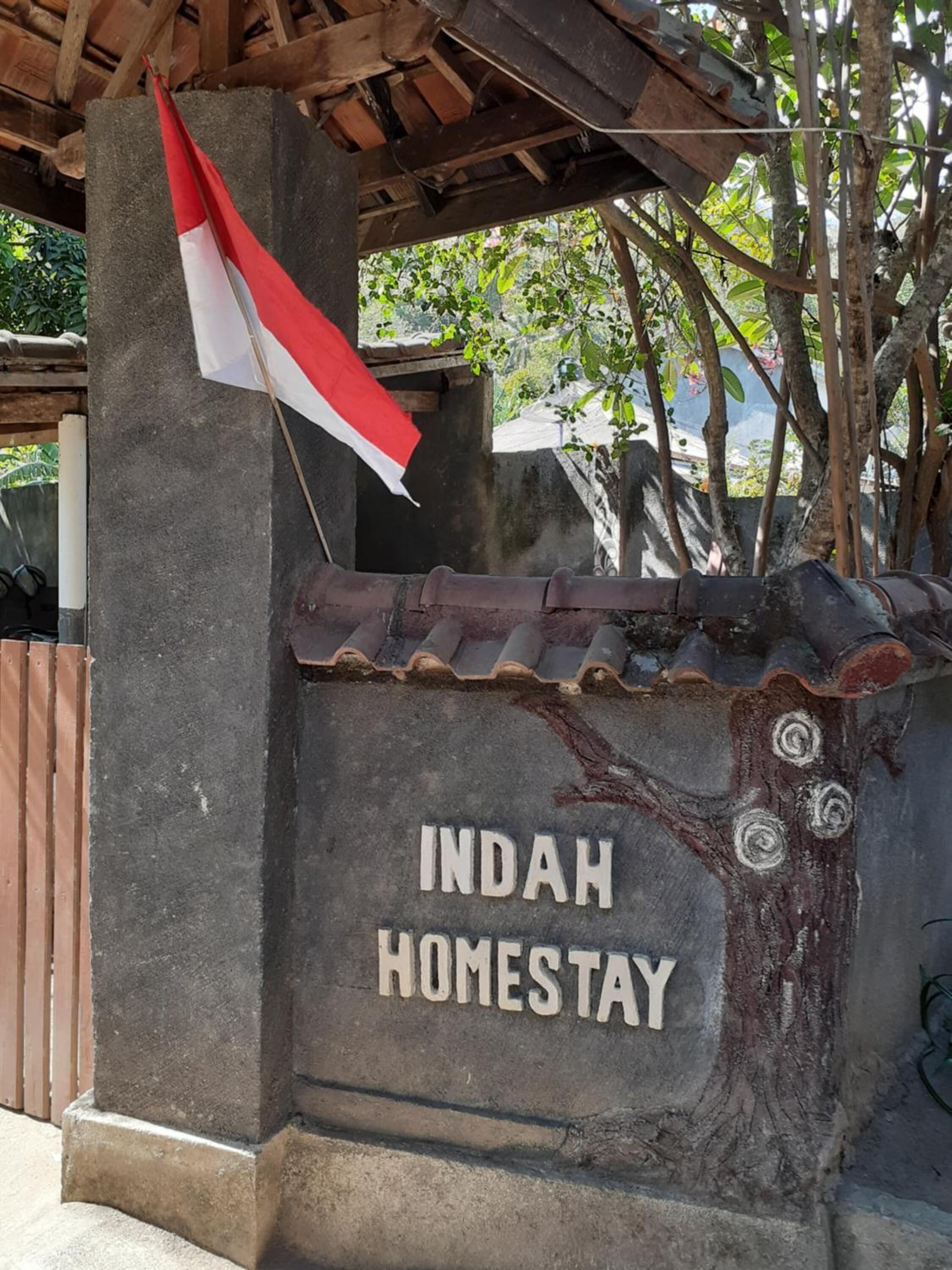 A stone sign with "INDAH HOMESTAY" written on it, a flag, and a tree with a tiled roof structure.