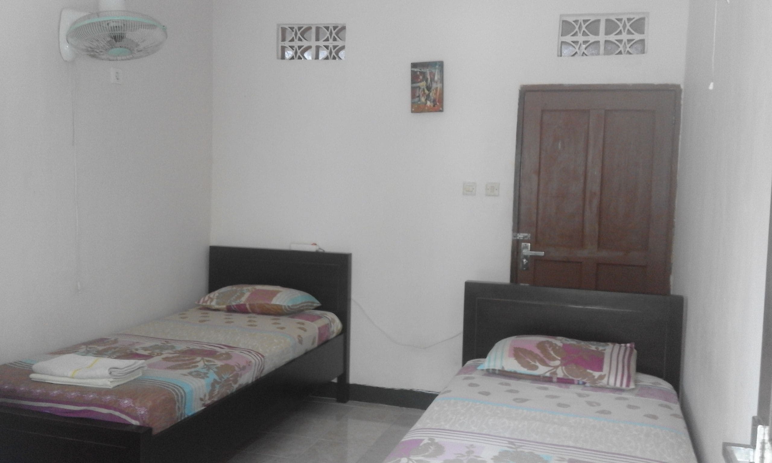 A small, simple room with two single beds, a wooden door, and a wall-mounted light fixture.
