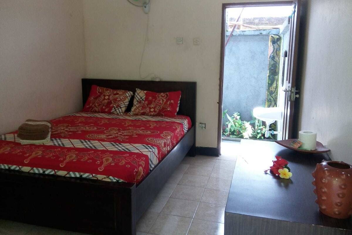 Une chambre à coucher avec un lit, une petite table sur laquelle se trouve un vase et des fleurs, et une porte donnant sur un espace extérieur.