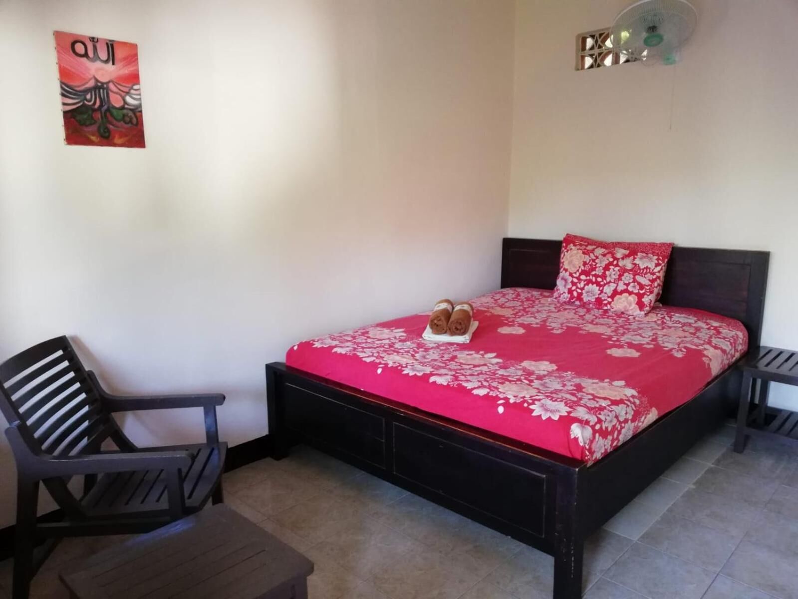 A bedroom with a bed, a chair, a small table, a wall decoration, and a ceiling fan.