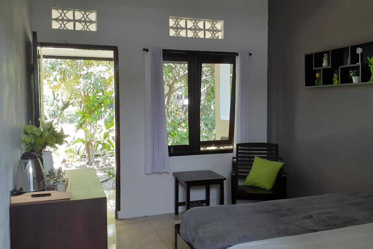 Une pièce comprenant un lit, une petite table, une chaise et une étagère ornée d’objets décoratifs. Il y a deux fenêtres avec des rideaux, et la vue donne sur un espace verdoyant.