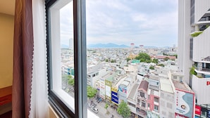 Vista urbana pela janela, mostrando edifícios, ruas e montanhas ao longe.