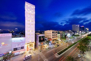 A multi-story hotel with a prominent vertical structure, a wide street with vehicles, and illuminated signage. - Mercury Boutique Hotel (Da Nang)