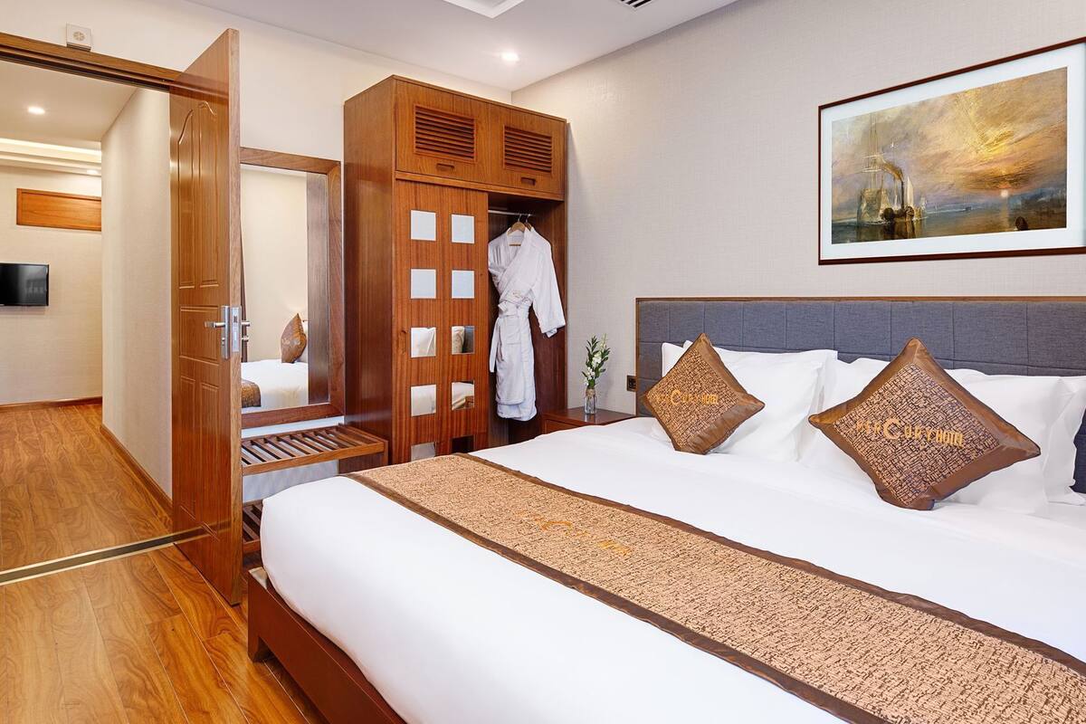A hotel room with a bed, a wooden wardrobe, a robe, a painting, and a mirror.