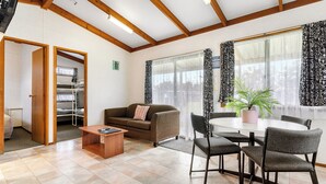 A modern living room with a sofa, coffee table, and dining area with chairs. There is a window with curtains, a plant on the table, and a door leading to another room. - Port Waikato Holiday Park (Port Waikato)