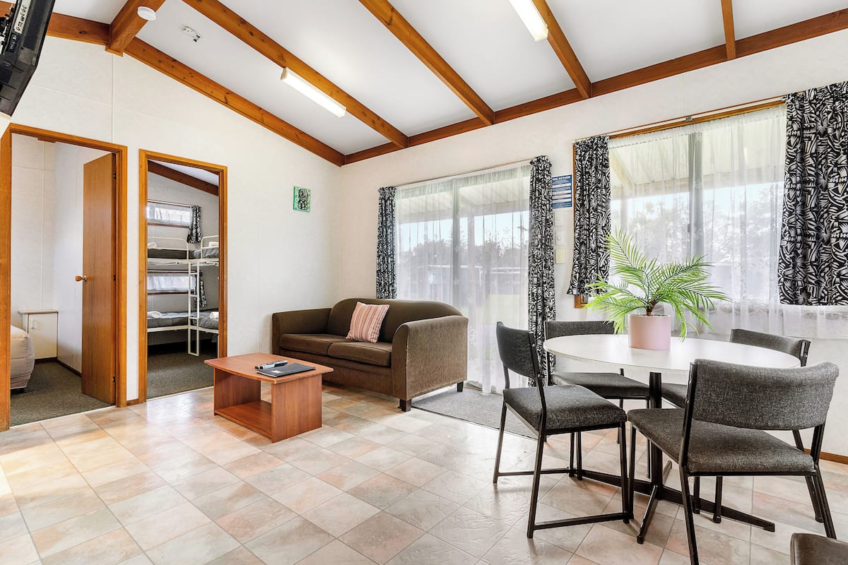 Een moderne woonkamer met een bank, salontafel en een eethoek met stoelen. Er is een raam met gordijnen, een plant op de tafel en een deur naar een andere kamer.
