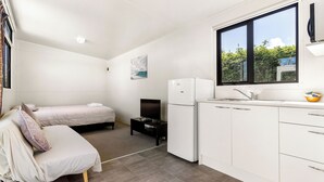A compact, modern room with a bed, a small kitchenette, and a window providing a view of greenery outside. - Port Waikato Holiday Park (Port Waikato)