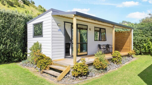 A modern single-story house with a covered porch, a small deck, and a landscaped garden with shrubs and a wooden bench. - Port Waikato Holiday Park (Port Waikato)